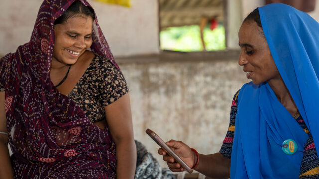 One lady in a maroon sari talking to a lady in blue sari, holding a smartphone, sitting in a white room.